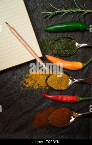 Page de l'ordinateur portable pour une recette et le crayon et les épices orientales dans les poivrons sur les cuillères métalliques dans le bac noir style Asiatique Banque D'Images