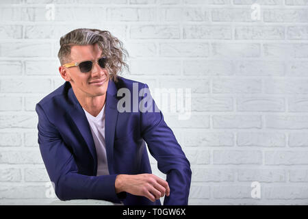 Homme élégant avec de longs cheveux bouclés dans lunettes assis sur une chaise sur fond blanc Banque D'Images