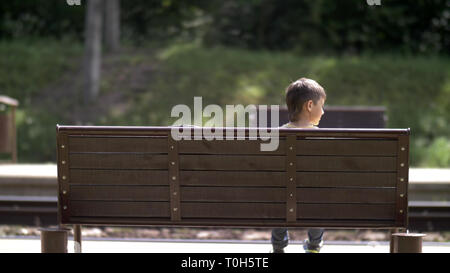 Garçon est assis sur un banc à la gare et attend que le train arrière à l'extérieur Banque D'Images