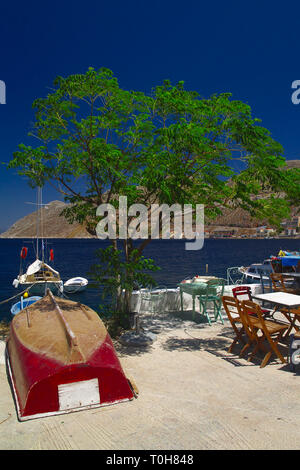 Un coup d'été de l'île de Symi en Grèce. Vue depuis un café avec tables chaises bateaux mer cristalline et maisons colorées. Banque D'Images