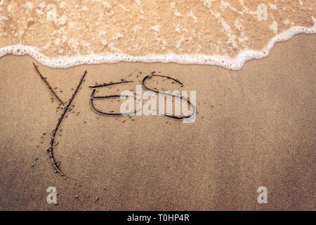Le mot 'oui' à la main sur le sable d'une plage au coucher du soleil avec des vagues sur le rivage Banque D'Images