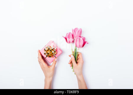 Woman's hands holding petite boîte cadeau rose avec archet d'or et bouquet de fleurs tulipe sur fond blanc, vue du dessus. Télévision lay concept minimal pour fem Banque D'Images