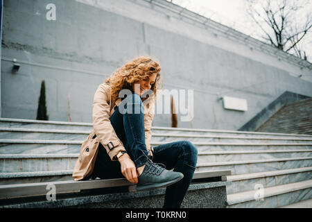 Happy young woman wearing trench-coat beige et bleu jeans assis sur des escaliers en pierre gris réglage de ses baskets. Banque D'Images