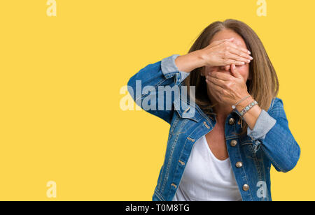 Belle femme d'âge moyen portant veste en jean décontracté sur fond isolé couvrant les yeux et la bouche avec les mains, surpris et choqué. La clandestinité emot Banque D'Images