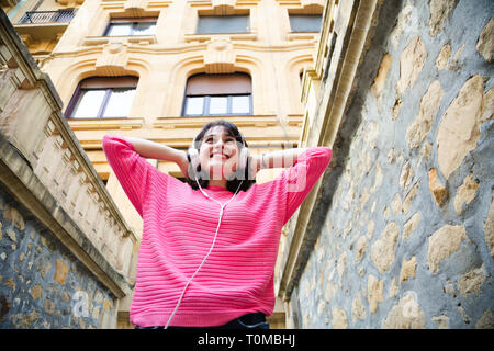 Happy young woman in pink sweater à écouter de la musique dans la rue. Low angle view Banque D'Images