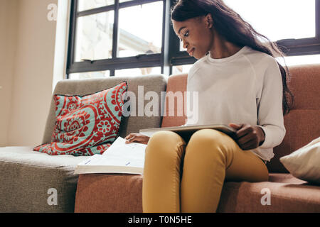 Jeune fille africaine assise sur un canapé avec un tablet pc la lecture d'un livre. Jeune étudiant étranger à l'intérieur à College campus. Banque D'Images