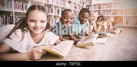 Les enfants de l'école sourire lying on floor reading book in library Banque D'Images