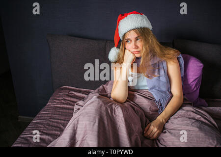 Jeune femme malade est assis sur lit et regarde droit. Elle s'ennuie. Femme détient les mains sous le menton. Elle a Christmas hat sur la tête. Banque D'Images