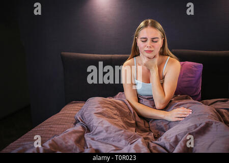 Photo de blonde s'assoit sur le lit et est titulaire d'une main sur la gorge. Elle a mal de là. Garde modèle les yeux fermés. Elle est seule dans la chambre. Banque D'Images