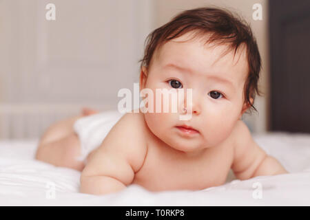 Portrait of cute adorable Asian mixed race baby girl 4 mois couché sur le ventre sur le lit dans la chambre à huis clos. L'émotion naturelle visage expressi Banque D'Images