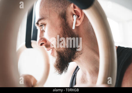 Élèvent dans les anneaux de gymnastique athlète masculin barbu portant un casque sans fil dans la salle de sport. Smiling fit man holding gymnast rings et lo Banque D'Images