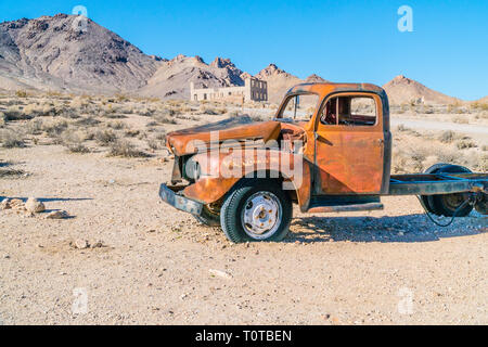 Un vieux camion rouge naufragé abandonné dans l'avant-plan et les ruines d'un bâtiment dans la ville fantôme de rhyolite, Nevada dans l'arrière-plan sur le s Banque D'Images