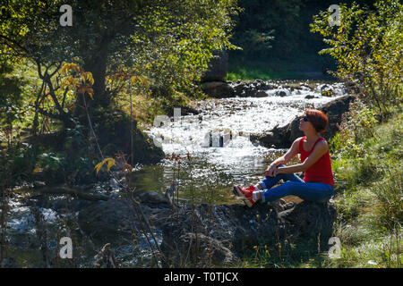 Jeune femme assise sur une pierre près d'un petit ruisseau sur une chaude journée d'automne Banque D'Images