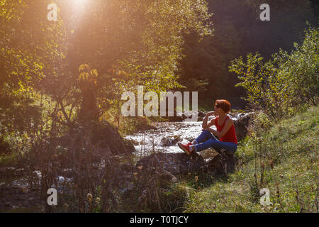 Jeune femme assise sur une pierre près d'un petit ruisseau sur une journée ensoleillée d'automne Banque D'Images