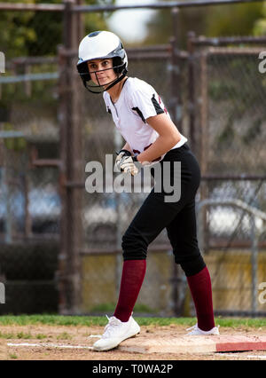 L'adolescence féminine de softball player en uniforme noir et blanc montrant de grands yeux à l'intérieur de son casque pendant le première base. Banque D'Images