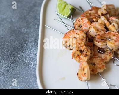 Crevettes épicées grillées sur des brochettes de métal avec le persil et le jus de lime ovale sur plaque blanche sur fond gris. Banque D'Images