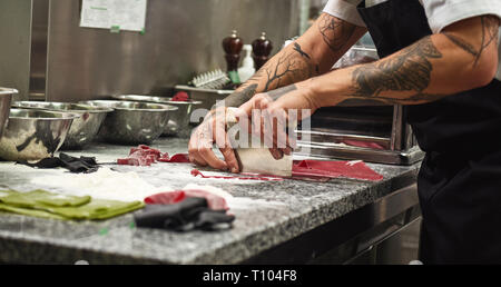 Faire des pâtes. La photo en gros plan de mains du chef avec des tatouages couper la pâte sur la table avec de la farine pour des pâtes dans la cuisine du restaurant Banque D'Images