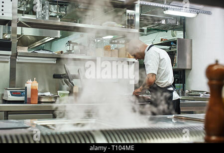 Journée bien remplie. Vue arrière de l'homme dans la coupe d'un chef de l'aire de la viande tout en se tenant dans une cuisine de restaurant. Cuisson Banque D'Images