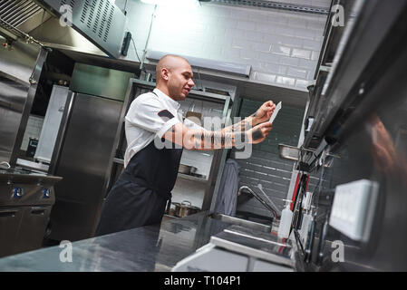 Quel est le prochain... Beau male chef chauve avec des tatouages sur ses bras à la recherche d'ordre liste dans un restaurant de cuisine. Concept de cuisine Banque D'Images