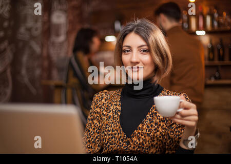 Freelancer blonde tenant une tasse de café tout en travaillant sur son ordinateur portable dans un pub. Banque D'Images