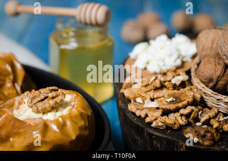 Pommes au four avec du fromage cottage et les écrous se trouvent dans un plat de cuisson noir sur une table en bois bleu à côté d'un pot de miel et une planche en bois sur laquelle les noix li Banque D'Images