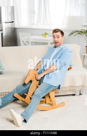 Bel homme excité à la caméra et de l'équitation à cheval à bascule en bois à la maison Banque D'Images