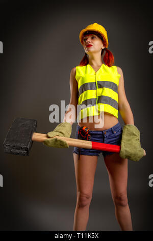 Une fille dans un casque jaune construction mitaines en shorts tenant un énorme marteau sur fond sombre Banque D'Images