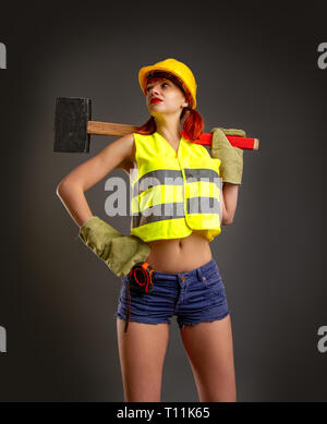 Une fille dans un casque jaune construction mitaines en shorts tenant un énorme marteau sur fond sombre Banque D'Images