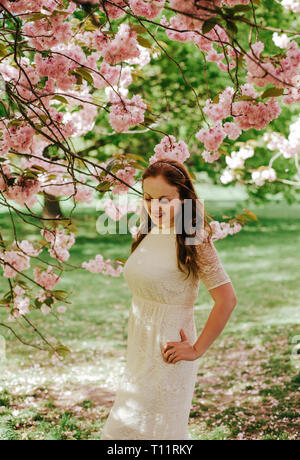 Belle brunette mignon fille mariée en dentelle crème, à décor sur les cheveux, près de l'arbre à fleurs roses fleurs, jardin, parc de Greenwich Banque D'Images