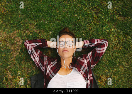 Top View of young woman wearing eyeglasses écoutez de la musique avec des écouteurs. Female student lying on grass avec les mains derrière la tête et de détente à col Banque D'Images