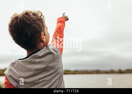 Vue arrière d'un enfant debout près du lac et montrant quelque chose à quelqu'un. Close up vue arrière d'un garçon pointant son doigt vers le ciel. Banque D'Images