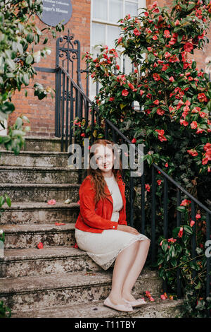 Belle jeune femme en robe en dentelle crème et veston orange assis sur des escaliers à l'extérieur de maison près de camelia fleurs rouges, smiling Banque D'Images
