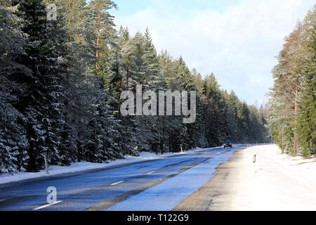 D'un paysage humide, route de campagne entourée de forêt avec l'approche de voiture au loin sur une journée ensoleillée d'hiver. Marttila, Finlande. Banque D'Images