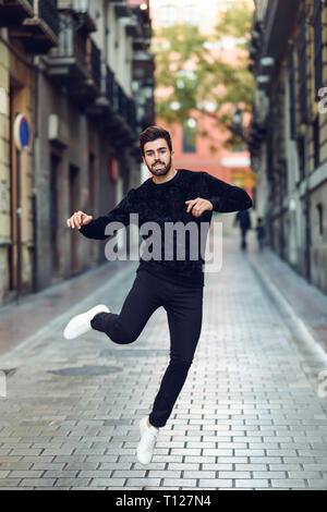 Jeune homme barbu qui saute dans l'arrière-plan urbain à bras ouverts portant des vêtements décontractés. Banque D'Images