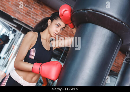 Young woman wearing boxing gloves in gym debout appuyée sur punching bag essuyant la sueur fatigue respiration Banque D'Images