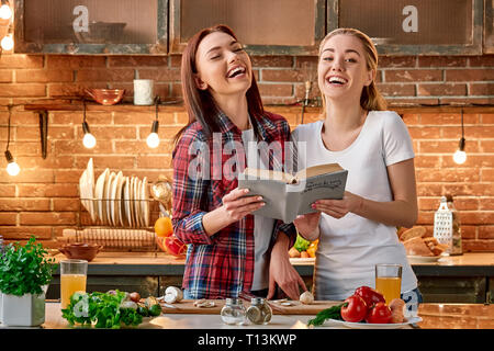 Portrait de deux femmes attrayants, s'amuser, tout en préparant la salade. Ils sont pleinement impliqués dans le processus. Fille blonde en T-shirt blanc et son ami brun en chemise à carreaux à la recherche de recettes santé dans un livre et de rire. Ils décident de cuisiner aujourd'hui. Vue avant Banque D'Images