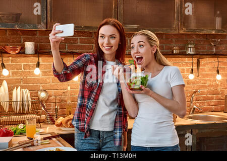 Portrait de deux femmes gaies, avoir du plaisir, le temps de préparer une salade de légumes. Dark-haired girl wearing chemise à carreaux est prenant, alors que sa blonde selfies ami en T-shirt blanc est titulaire d'une salade cuite et pose pour une photo Banque D'Images