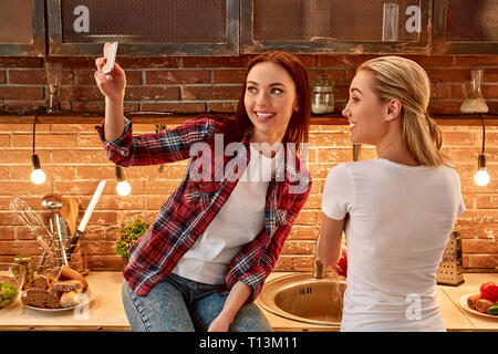 Portrait de deux femmes attrayants, s'amuser, tout en préparant une salade de légumes. Blonde girl in white T-shirt est avec son dos à la caméra et se lave les légumes dans l'évier et son amie portant chemise à carreaux prend tout en étant assis sur le selfies d'examen. Vue avant Banque D'Images