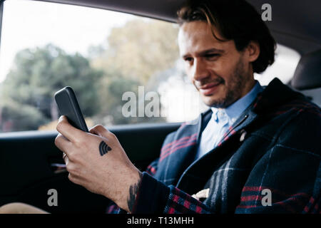 Smiling man sitting on siège arrière d'une voiture à l'aide de téléphone cellulaire Banque D'Images