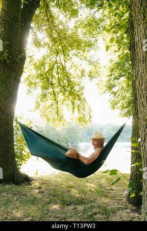 Senior man wearing straw hat relaxing in hammock au lakeshore reading book Banque D'Images