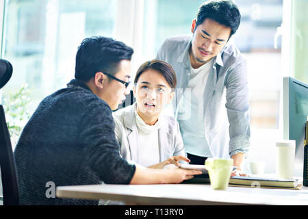 Trois jeunes dirigeants de sociétés asiatiques meeting in office discuter affaires using digital tablet. Banque D'Images