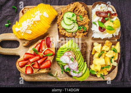 Différents types de sandwichs à la mangue, fraise, tofu pate, avocat, pommes de terre et de la ricotta sur un fond sombre. Banque D'Images