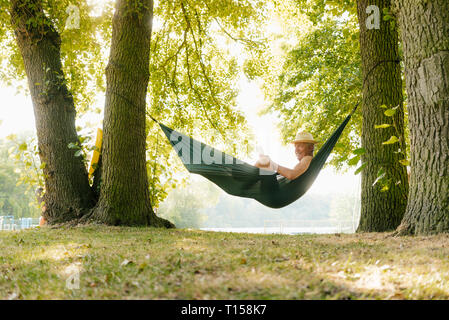 Senior man wearing straw hat relaxing in hammock au lakeshore reading book Banque D'Images