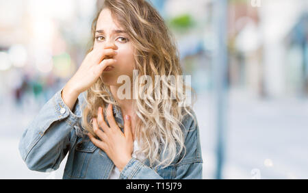 Très belle jeune femme blonde wearing denim veste sur fond isolé humer un stinky et dégoûtant, odeur insupportable, apnée Banque D'Images