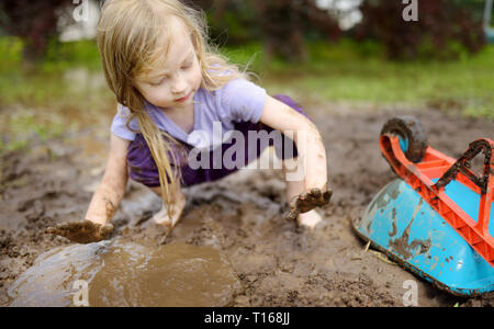 Funny little girl playing dans une grande flaque de boue humide par beau jour d'été. Enfant de se salir en creusant dans le sol boueux. Jeux en plein air. malpropre Banque D'Images