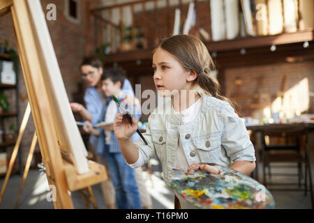 Vue de côté de l'accent portrait petite fille photo peinture sur chevalet dans la classe d'art et la palette holding, copy space Banque D'Images