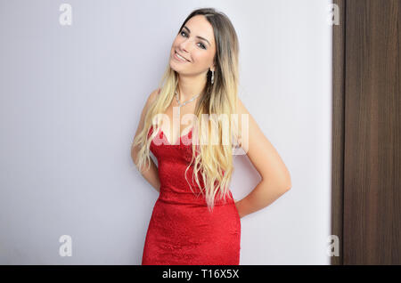 Beau poli model wearing red dress, debout contre le mur. Les jeunes, fit woman posing in studio avec fond blanc. Banque D'Images