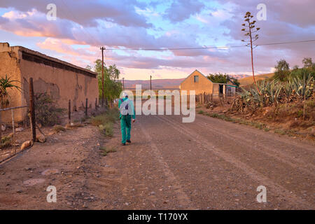 Karoo route de terre menant à une courbure dans la lumière du matin. L'homme en vert dans l'ensemble par farm house vu de retour au travail à pied Banque D'Images