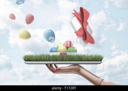Vue latérale du woman's hand holding tray patch owith de pelouse verte avec open gift box en haut à partir de laquelle les ballons colorés sont flying out against cloudy Banque D'Images