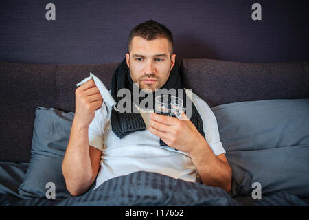 Distrait les jeunes homme malade est assis sur le lit avec couverts faible du corps. Il est titulaire d'une serviette blanche et de verre sur l'eau. Il y a aussi d'écharpe autour de son cou Banque D'Images